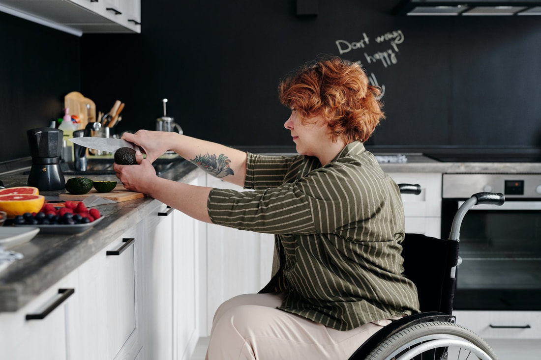 Femme-presenting person in a wheelchair cutting an avocado. In the background, a message in chalk reads 